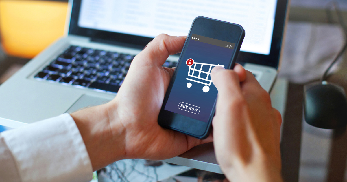 Two hands hold a mobile phone that displays a shopping cart, suggesting shopping on the go. A laptop computer is blurred in the background.